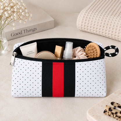 Neceser de neopreno blanco con puntos negros y franja central roja, abierto y con productos de cuidado personal.