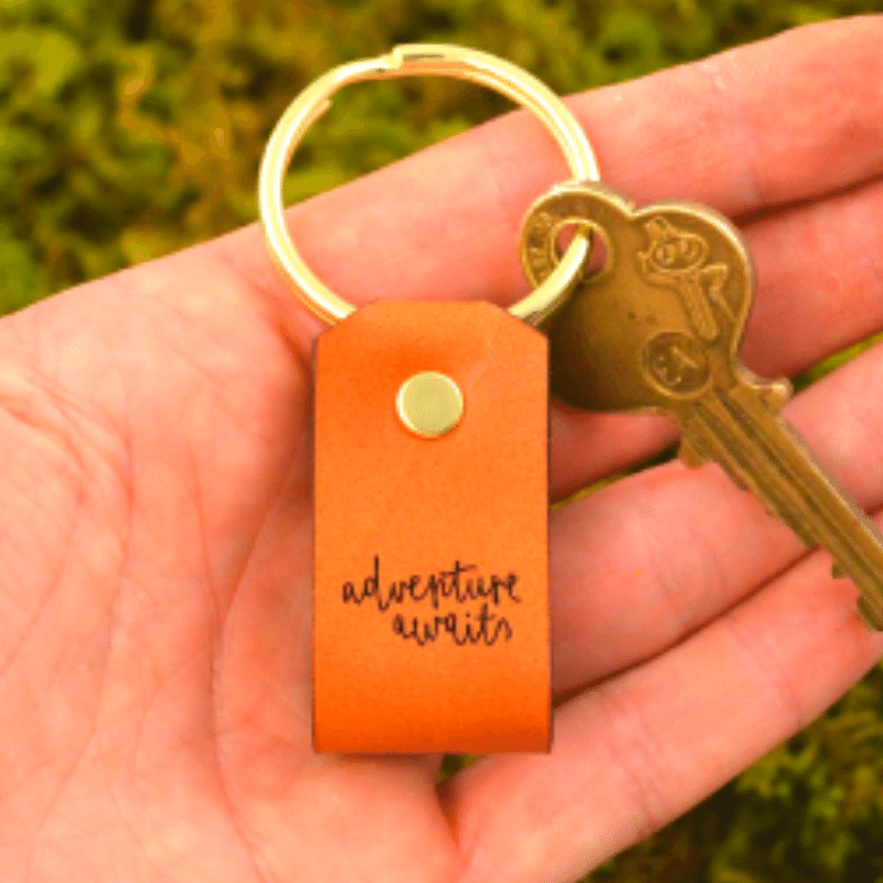 Llavero de cuero con mensaje “Adventures Awaits”, anillo de latón y diseño elegante. Ideal como regalo con significado y estilo.